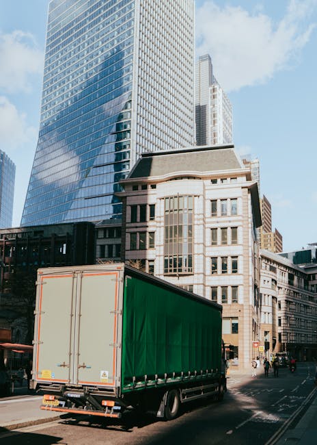 A large delivery truck with a green cargo area is parked on the street near the entrance of a building in central London, under a clear sky. Behind the truck, there are tall modern skyscrapers with reflective glass facades and a shorter, older-style building with stone and brickwork. The truck is positioned adjacent to the building's curbside, where a small set of stairs leads up to the entrance. The area appears busy, with pedestrians walking along the sidewalk and some people near the building. The truck is part of a house or office relocation service, and the scene suggests a furniture transport operation involving loading or unloading near an access point on Old Kent Road, with visible urban infrastructure supporting the logistics of a home or commercial move. Man and Van Southwark may handle such removals, including navigating access challenges in this area.