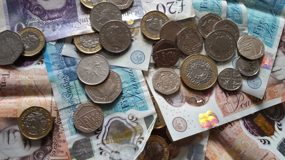 A close-up image displaying a scattered assortment of British currency, including various coins such as £1, 50p, 20p, 10p, and smaller denominations, alongside multiple banknotes of different denomina