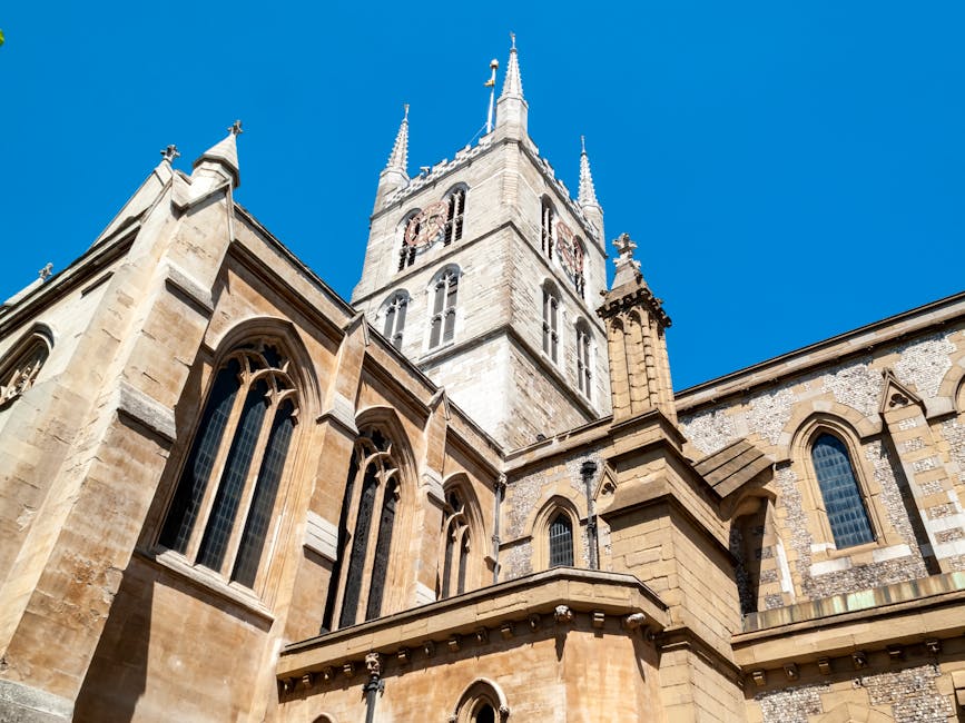 A low-angle view of a historic cathedral with beige stone walls and pointed Gothic-style arched windows, set against a bright blue sky. The cathedral features intricate architectural details including tall spires and buttresses. A wooden step ladder and some construction scaffolding are partially visible along one side of the building. The scene captures a moment of exterior work possibly related to building maintenance or restoration. This type of setting can be associated with relocation or preservation projects involving historic property, which may be coordinated by companies like Man and Van Southwark, often involved in house removals or furniture transport for homes near such landmarks.