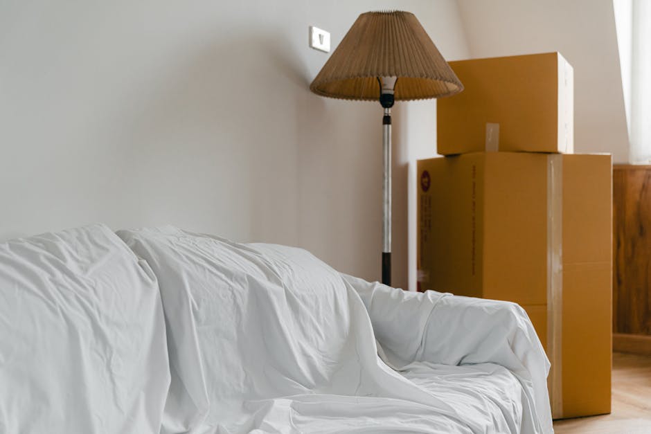 Interior scene showing a living room corner with a white sofa covered by a protective sheet, positioned near a wall with a light switch and a standing floor lamp with a beige lampshade. To the right, there are three cardboard moving boxes stacked, with two boxes on the bottom and one on top, all sealed and ready for packing or transport. The floor appears to be wooden, and natural light enters from an unseen window, illuminating the space. The setting suggests a home during a packing or moving process, highlighting furniture, packing materials, and the environment involved in home relocation activities, as provided by Man and Van Southwark.