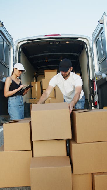 Two individuals are engaged in a home relocation process involving furniture transport and packing within a large, open van with its rear doors wide open. The man, dressed in a white shirt and black cap, is seen lifting and stacking several medium-sized cardboard boxes in front of the van, ensuring the boxes are securely positioned for transportation. The woman, wearing a baseball cap and casual attire, stands nearby holding a clipboard, possibly overseeing or documenting the moving and packing process. Inside the van, additional cardboard boxes filled with household items are visible, neatly stacked to optimize space and protect the items during transit. The van is parked outdoors, with a clear sky overhead, and the scene is well-lit by natural daylight, illustrating a professional move involving careful handling of packaging materials such as cardboard boxes, and the use of equipment like trolleys or straps might be implied. This image reflects typical steps in a moving service provided by Man and Van Southwark, focusing on the loading process during a house removal or relocation effort, aligned with the topic of avoiding common packing mistakes in household moves.