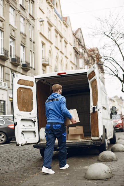 Two movers from Man and Van Southwark are loading cardboard boxes into the back of a large white van parked outside a modern building with glass windows. One mover, wearing gloves and a blue jacket, is passing a box to another person inside the van, who is wearing a black jacket, white sneakers, and a cap, while taking notes or checking a clipboard. Several boxes, some labeled with fragile stickers and packing symbols, are positioned on the pavement and near the van entrance. The interior of the van reveals additional boxes and packing materials, indicating a home relocation or furniture transport process. The scene is set outdoors in daylight, with the large van partially open, ready for loading or unloading, illustrating the logistics involved in packing and moving services provided by Man and Van Southwark.