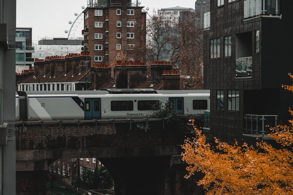 A Thameslink train with a grey and blue exterior is seen passing over a brick railway bridge during daylight, with residential buildings and a modern black glass apartment block visible in the backgro