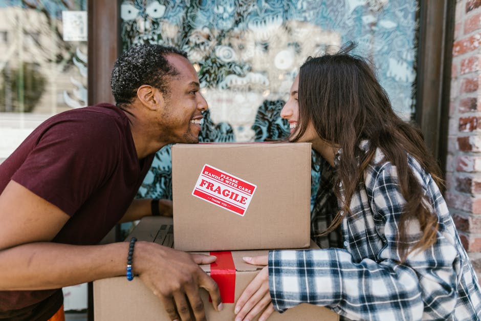 An image displaying a man and a woman engaged in a home relocation process outside a property, with both individuals smiling and holding cardboard boxes. The man, with dark curly hair and wearing a maroon T-shirt, is holding the top of a cardboard box marked 'Fragile' with a red and white sticker, while the woman, with long brown hair and dressed in a blue and white checked shirt, is supporting a larger box below. The boxes are wrapped in brown packaging tape. In the background, there is a section of a building facade with a large window featuring decorative patterns, and the setting appears well-lit with natural daylight. The scene captures the loading or unpacking stage of furniture transport or packing and moving services, with visible emphasis on careful handling of fragile items, typical of professional removals performed by companies like Man and Van Southwark.