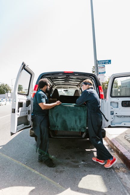 Two removal professionals from Man and Van Southwark are loading a large, dark green covered piece of furniture into the back of a white van parked on a street. The open rear doors reveal the vehicle's spacious interior, which is empty except for the furniture being carefully positioned inside. One worker, wearing a blue uniform, is holding the furniture steady, while the other, dressed in a dark jacket and red running shoes, aids in the lifting process. The scene takes place outdoors on a bright day, with a clear sky and a street sign visible in the background. This image captures the careful handling and strategic loading involved in home relocation and furniture transport, highlighting the significance of proper packing and moving logistics provided by [COMPANY_NAME], which specialises in removals and house moves in the Southwark area.