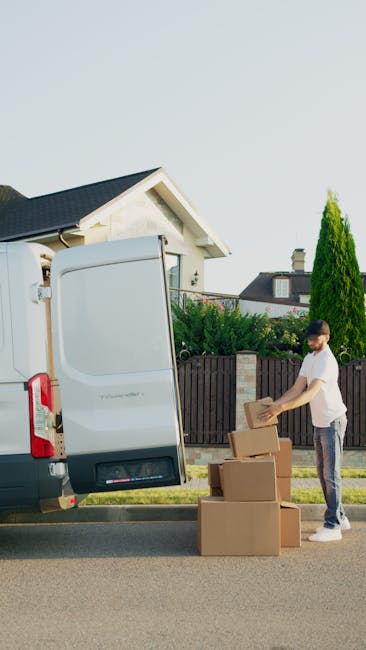An image displaying a man and a woman engaged in a home relocation process outside a property, with both individuals smiling and holding cardboard boxes. The man, with dark curly hair and wearing a maroon T-shirt, is holding the top of a cardboard box marked 'Fragile' with a red and white sticker, while the woman, with long brown hair and dressed in a blue and white checked shirt, is supporting a larger box below. The boxes are wrapped in brown packaging tape. In the background, there is a section of a building facade with a large window featuring decorative patterns, and the setting appears well-lit with natural daylight. The scene captures the loading or unpacking stage of furniture transport or packing and moving services, with visible emphasis on careful handling of fragile items, typical of professional removals performed by companies like Man and Van Southwark.
