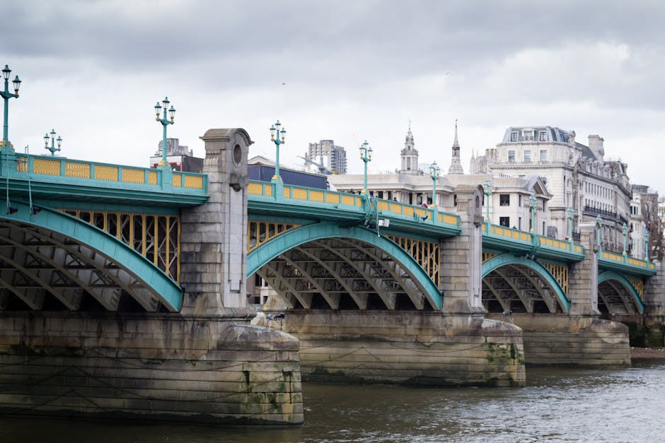 A detailed view of a large, historic metal and stone bridge spanning a river in an urban setting, featuring turquoise and yellow railings, ornate lamp posts, and stone piers, with high-rise and classical-style buildings visible in the background under an overcast sky. The image captures the bridge during a home relocation or moving process, with the lower part of the bridge and riverbank in clear view, providing context for vehicle parking and logistical planning for removals. This setting illustrates the typical environment where Man and Van Southwark operates, highlighting the importance of careful planning during furniture transport across city infrastructure such as this bridge, with no visible furniture, boxes, or moving equipment present.