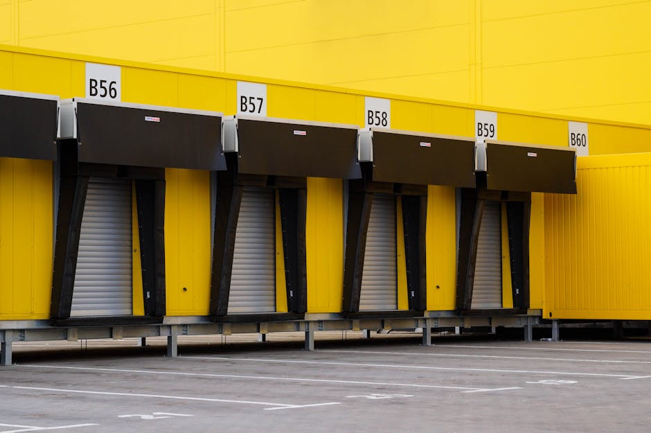 A row of four yellow and black loading dock bays at a warehouse or storage facility in Southwark, SE1, with each bay numbered B56, B57, B58, and B59. The bays have black retractable dock levelers or lifts in a closed position beneath black canopies. The loading area is an outdoor paved space with painted white lines for parking or vehicle positioning, and the background features bright yellow exterior walls supporting the loading bays. This setting illustrates a typical commercial property prepared for furniture or equipment transport related to home relocation or moving services, as managed by Man and Van Southwark, highlighting the logistics of loading and unloading during moving operations.