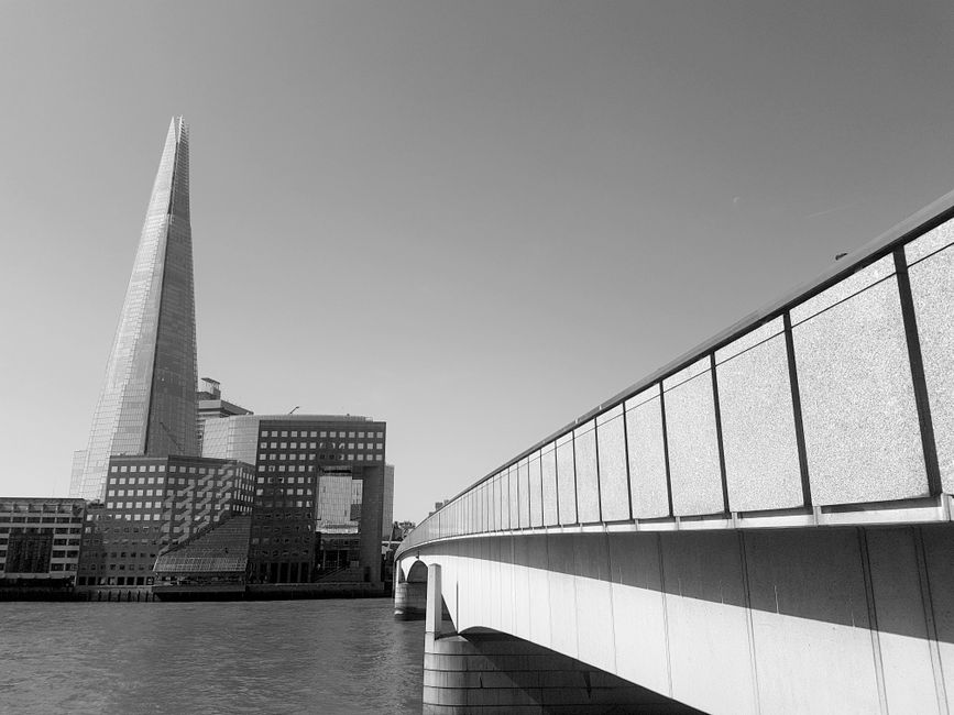 A black and white photograph depicting a cityscape view from a riverside location in London, featuring the towering Shard building on the left and several modern office buildings with grid-patterned windows. In the foreground, a concrete bridge with a metal railing runs horizontally across the image, casting shadows on its underside. The scene is illuminated by natural light, highlighting the architectural details and the reflective water below, capturing the urban environment associated with house removals and moving logistics in Southwark. The image exemplifies an area where loading bay rules and fines may apply for residential and commercial relocations, as managed by Man and Van Southwark, in the context of home relocation, furniture transport, and packing and moving operations.