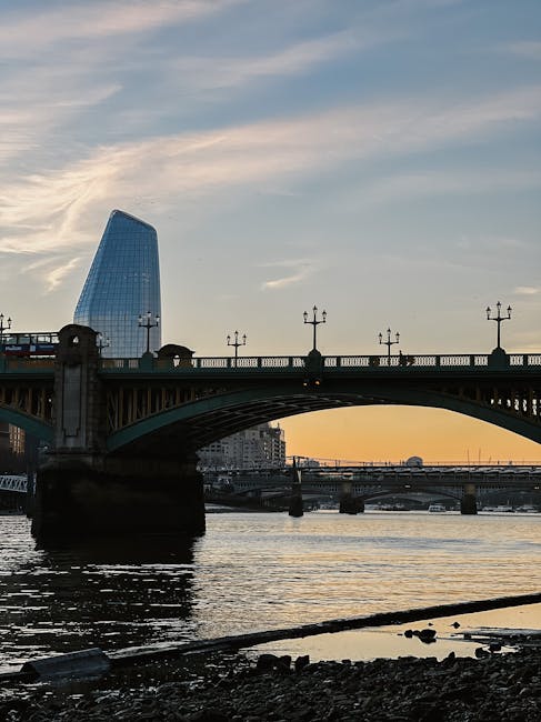 A view of a large steel bridge spanning a river during sunset, with decorative lampposts along the bridge's balustrade. In the background, there is a modern, uniquely shaped glass skyscraper with a curved, angular design, reflecting the sky and nearby buildings. The river below shows gentle ripples, and the shoreline in the foreground includes rocks and a partially submerged rope or cable. The scene is well-lit with natural evening light, capturing the urban landscape near London Bridge in Southwark, relevant to home relocation or furniture transport through city environments, as experienced in professional removals services by Man and Van Southwark.