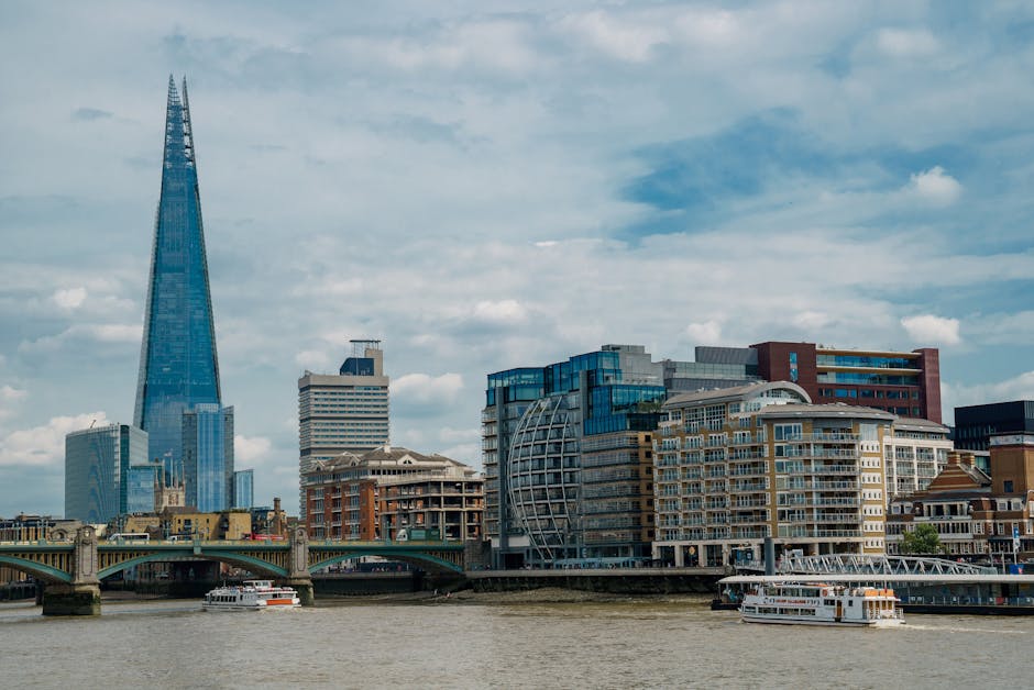 A cityscape view of Southwark in London showing modern high-rise buildings and historic structures along the River Thames. The Shard, a tall, glass skyscraper, dominates the skyline on the left side of the image. In the foreground, there is a stone bridge crossing the river, with a white passenger boat passing underneath. To the right, residential and commercial buildings with balconies and large windows line the riverbank. The sky above features scattered clouds with patches of blue, and the overall scene suggests daytime with natural daylight. This setting is relevant to house removals and furniture transport, illustrating urban logistics for home relocation services. The image is associated with the services of Man and Van Southwark, as seen on the webpage titled 'Tower Bridge Road man and van removals Southwark'.