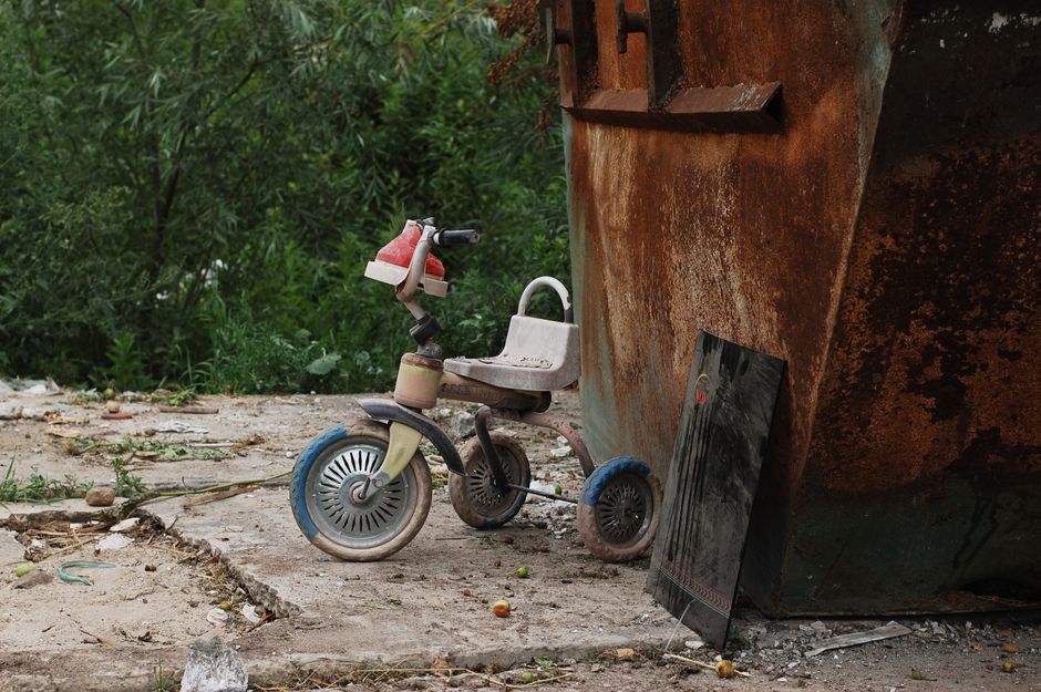 The image shows a small, weathered children's tricycle with a pink and white frame, black handlebars, and large, circular plastic wheels, positioned on a cracked concrete surface outdoors. The tricycle appears old and worn, with visible dirt and grime, and is situated next to an old, rusted metal boat hull that is leaning against a wall or structure. Behind the boat, there is dense greenery with various trees and bushes, suggesting a backyard or outdoor storage area. Nearby, a rectangular piece of black material, possibly a blackboard or panel, is propped against the boat. The scene captures a moment of home relocation or decluttering, with objects that may be being considered for removal or disposal, aligning with services related to house removals or bulk waste management, as indicated on the webpage about skips and bulky waste rules for Southwark Council. The overall environment appears untidy and in need of organizing, consistent with the context of moving or clearing out a property, and the image is lit with natural daylight.