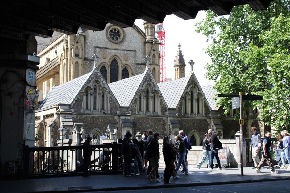 A group of pedestrians walking along a city street under a bridge, with a historic stone church visible in the background. The church features arched windows, detailed stonework, and multiple spires. The scene is shaded by trees on the right side, and a variety of people, some in groups and others alone, are moving along the pavement. The image captures an urban environment with a mix of historic architecture and active foot traffic, relevant to the process of home relocation and moving logistics managed by companies like Man and Van Southwark. The lighting suggests daytime, with sunlight filtering through the trees and creating contrasting shadows. The area appears busy, reflecting themes of packing, furniture transport, and the logistical aspects of house removals, particularly in a historic setting near Southwark.